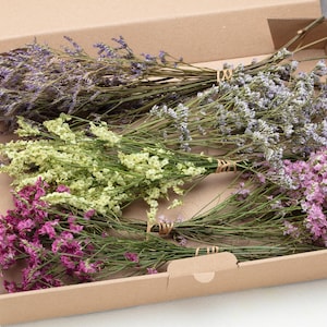 Könnte beinhalten: Ein brauner Karton mit mehreren Bündeln getrockneter Blumen. Die Blumen sind in Lila, Rosa und Grün gehalten und mit Bindfäden zusammengebunden. Die Schachtel ist offen und zeigt die farbenfrohe Anordnung.