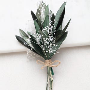 Könnte beinhalten: Ein kleines Bouquet aus dunkelgrünen Olivenblättern und zartem weißen Schleierkraut, gebunden mit Bindfaden. Das Arrangement befindet sich vor einem weißen Hintergrund und erzeugt eine natürliche und elegante Ästhetik. Ideal für Hochzeiten oder besondere Anlässe.