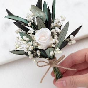 Op de afbeelding: Een corsage met een enkele witte roos omringd door donkergroene olijfbladeren en kleine witte gipskruidbloemen. De stelen zijn samengebonden en vastgezet met een dun, natuurlijk bruin touwtje dat in een strik is geknoopt.