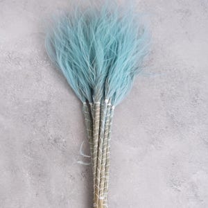 May include: A bunch of light blue dyed feathers, bound together with natural-coloured stems wrapped in white thread. The feathers have a soft, fluffy texture and a delicate look. The background is a light grey, textured surface.