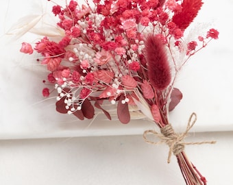 May include: A bouquet of dried flowers in shades of pink and red, tied with twine. The bouquet includes small pink flowers, red and white feathery stems, and dried eucalyptus leaves.
