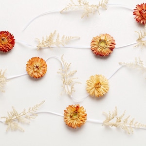 Peut inclure: Une guirlande de ficelle blanche avec des fleurs séchées dans des tons d'orange et de jaune. Les fleurs sont disposées en cercle et espacées uniformément.