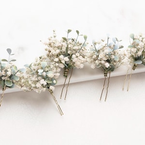 Può includere: Set di cinque forcine per capelli floreali bianche e verdi. Ogni forcina presenta un piccolo bouquet di gipsofila bianca e fogliame verde.