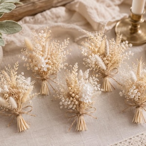 Puede incluir: Cinco peque&ntilde;os ramos de flores y hierbas secas en tonos crema, beige y blanco. Cada manojo est&aacute; atado en la base con un lazo fino de cuerda de color natural. Los arreglos presentan una mezcla de texturas que incluyen colas de conejo esponjosas, paniculata y peque&ntilde;as flores secas.