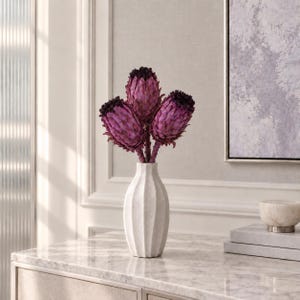 Könnte beinhalten: Eine weiße Keramikvase mit vertikaler Rippenstruktur enthält drei tiefviolette Protea-Blüten. Die Blüten haben dunkle, strukturierte Mitten und geschichtete, schuppenartige Blütenblätter, die einen Kontrast zur glatten, matten weißen Oberfläche der Vase bilden.