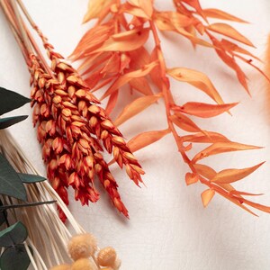 May include: Close-up of dried floral arrangement with orange and green hues. The arrangement features various textures, including fluffy, feathery, and spiky elements. The color palette includes shades of orange, green, and cream, creating a warm and natural aesthetic.