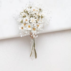 Op de afbeelding: Een klein boeket witte madeliefachtige bloemen met gele centra en delicate witte vulbloemen. De stelen zijn gebonden met touw en groene stelen. De opstelling staat tegen een witte achtergrond.