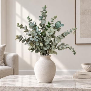 May include: A round, off-white ceramic vase with a speckled texture holds a large arrangement of green eucalyptus branches. The vase features a wide, bulbous body that tapers towards a narrow neck and a flared rim, displaying natural foliage.