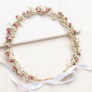 Puede incluir: Una corona floral con pequeñas rosas rosas, paniculata blanca y follaje verde. La corona circular está atada con una cinta de satén blanco. La corona está sobre un fondo blanco.