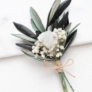 Könnte beinhalten: Ein Anstecker mit einer weißen Rose und kleinen weißen Blüten, akzentuiert durch Olivenblätter. Die Anordnung ist mit Bindfaden gebunden. Die Blätter sind dunkelgrün und schwarz.