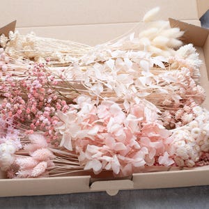 Op de afbeelding: Een kartonnen doos gevuld met een assortiment gedroogde bloemen in tinten roze, crème en beige. De opstelling omvat pluizige konijnenstaarten, delicate takjes en grotere bloemen, wat een zachte, natuurlijke uitstraling creëert.