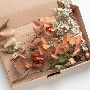 Peut inclure: Une boîte en carton remplie de fleurs et de feuillage séchés. Les fleurs sont un mélange de blanc, de rose et de brun. Le feuillage est un mélange de vert et de brun.