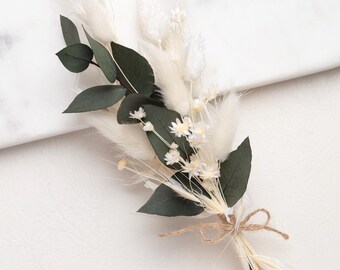 May include: A dried flower bouquet featuring white fluffy pampas grass, small white flowers, and dark green leaves. The bouquet is tied with twine and rests on a white surface. A natural, elegant floral arrangement.