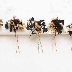 May include: Five hair pins with black and gold floral detailing. Each pin has a cluster of black and gold leaves and petals, attached to a thin, gold-coloured wire. The pins are arranged on a white surface, creating a decorative hair accessory.