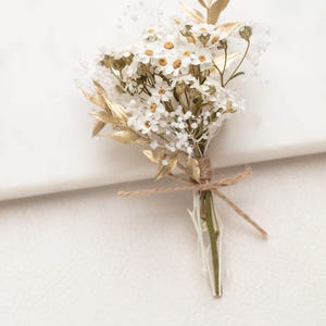Peut inclure: Un petit arrangement floral délicat composé de fleurs blanches à cœur jaune et de feuilles dorées. Les tiges sont enveloppées et liées avec de la ficelle. Il s'agit probablement d'une boutonnière ou d'un petit bouquet.