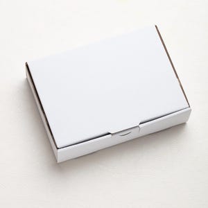 May include: A white, rectangular cardboard box with a closed lid. The box has a simple, clean design, with a smooth, matte finish. The edges of the box are a light brown color.