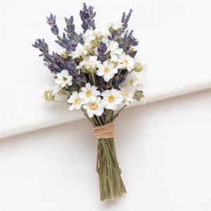 Könnte beinhalten: Ein kleiner Strauß aus getrockneten Blumen mit Lavendel und weißen Gänseblümchen. Die Stiele sind mit Bindfaden gebunden. Die Blumen liegen auf einer weißen Oberfläche.
