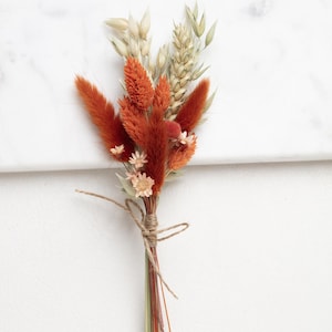 Op de afbeelding: Een klein boeket gedroogde bloemen in tinten oranje en beige, gebonden met touw. Het boeket bevat gedroogde grassen, kleine oranje bloemen en pluche oranje pluimen.
