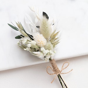 Könnte beinhalten: Ein Ansteckblumen-Arrangement aus getrockneten weißen und blassgrünen botanischen Elementen, darunter flauschige Hasenschwanzgräser, zartes Schleierkraut und schlanke Halme. Die Stiele sind mit natürlichem Jutegarn umwickelt und mit einer kleinen Schleife versehen.