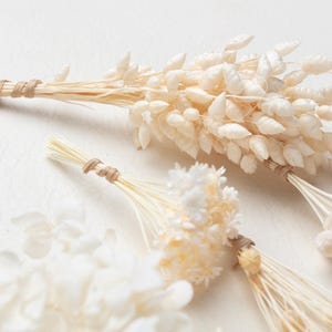 Puede incluir: Una colecci&oacute;n de ramos de flores secas con hierbas y peque&ntilde;as flores en tonos blanco roto y crema. Cada manojo est&aacute; cuidadosamente atado en la base de los tallos con un cordel fino de fibra natural de color marr&oacute;n claro, formando un conjunto bot&aacute;nico.
