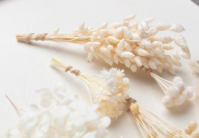 Puede incluir: Una colecci&oacute;n de ramos de flores secas con hierbas y peque&ntilde;as flores en tonos blanco roto y crema. Cada manojo est&aacute; cuidadosamente atado en la base de los tallos con un cordel fino de fibra natural de color marr&oacute;n claro, formando un conjunto bot&aacute;nico.