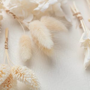 Puede incluir: Una colecci&oacute;n de elementos bot&aacute;nicos secos que incluye hierba de cola de conejo de color crema, p&eacute;talos de hortensia blancos preservados y peque&ntilde;os ramos de flores secas atados con un cordel fino de fibra natural.