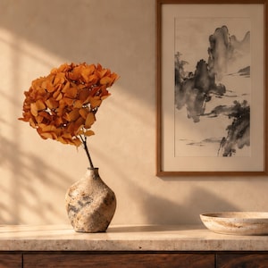 May include: A textured, speckled ceramic vase holds a cluster of vibrant orange hydrangea flowers. Beside it sits a shallow, circular stone bowl. On the wall hangs a framed black and white ink wash painting depicting a mountainous landscape scene.