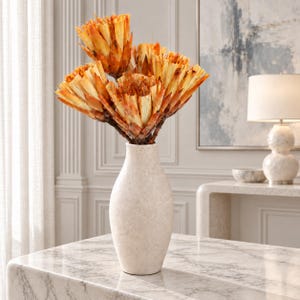 Könnte beinhalten: Eine hohe, cremeweiße Keramikvase mit gesprenkelter, matter Textur enthält einen Strauß getrockneter, goldorangefarbener Protea-Blüten. Die Blüten haben geschichtete, spitze Blätter mit dunkelbraunen Mitten und bilden ein strukturiertes Arrangement.