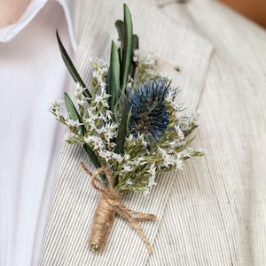 Könnte beinhalten: Ein kleiner Strauß getrockneter Blumen mit weißen Blüten und blauen Disteln, mit Schnur gebunden, an einem hellbraunen Leinenjackett befestigt.