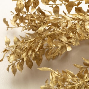 Può includere: Primo piano di una composizione botanica dorata. L'immagine presenta diversi rami con piccole foglie dettagliate, tutti rivestiti con una finitura oro metallizzato. Le foglie hanno un aspetto strutturato e la composizione è su uno sfondo neutro.