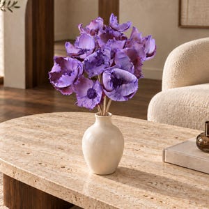 Könnte beinhalten: Ein Strauß aus leuchtend violetten Blumen mit gerüschten Blütenblättern und dunklen Mitten, arrangiert in einer glatten, cremeweißen Keramikvase. Die Blumen haben mehrere Blüten an dünnen, hellen Stielen und bilden ein dichtes, rundes Arrangement.
