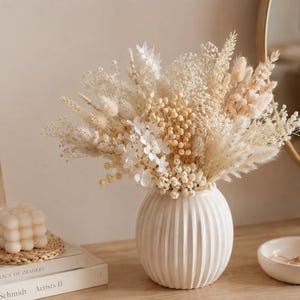 Puede incluir: Un jarr&oacute;n de cer&aacute;mica blanca con estr&iacute;as verticales contiene un denso arreglo de flores y hierbas secas en tonos crema, beige y blanco roto. El ramo incluye hierba de la pampa esponjosa, paniculata delicada y peque&ntilde;os racimos florales.