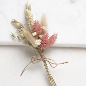 Könnte beinhalten: Ein kleiner Strauß getrockneter Blumen, die mit einer Schnur zusammengebunden sind. Der Strauß enthält rosa und weiße Blumen, getrocknete Gräser und Weizen.