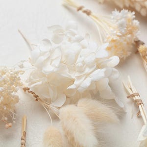 Puede incluir: Una colecci&oacute;n de elementos florales secos y preservados que incluye floretes de hortensia blancos, colas de conejo de color crema esponjosas y peque&ntilde;os racimos de delicada paniculata blanca, todos atados con cordel natural fino en los tallos.