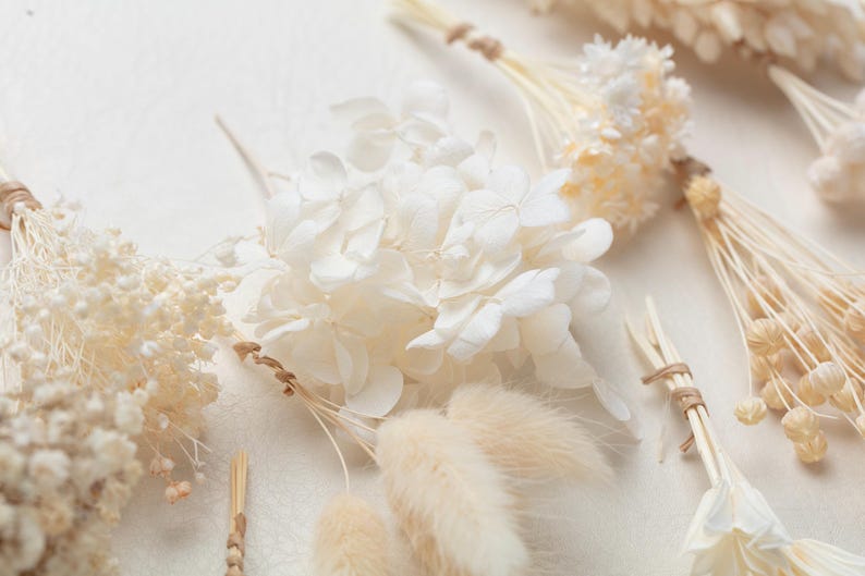 Puede incluir: Una colecci&oacute;n de elementos florales secos y preservados que incluye floretes de hortensia blancos, colas de conejo de color crema esponjosas y peque&ntilde;os racimos de delicada paniculata blanca, todos atados con cordel natural fino en los tallos.