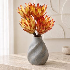 May include: A gray ceramic vase with a twisted, textured surface holds a bouquet of four large protea flowers. The flowers feature pointed petals that transition from deep red at the tips to bright yellow at the base, with dark centers.