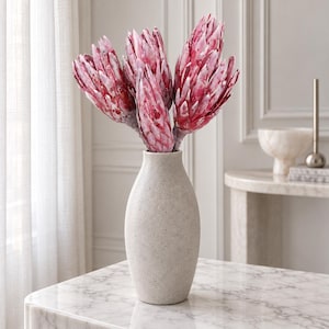 Könnte beinhalten: Eine hohe, hellbeige Vase aus strukturiertem Stein enthält einen Strauß aus fünf großen, leuchtend rosa Protea-Blüten. Die Blüten haben geschichtete, spitz zulaufende Blätter mit einer gesprenkelten Färbung in Magenta und Weiß.