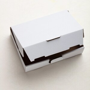 May include: A white cardboard box with a partially open lid. The box is rectangular and has brown edges visible where the lid is open. The box is sitting on a white surface.
