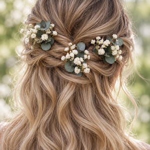 Könnte beinhalten: Drei Haarschmuck-Elemente bestehend aus Büscheln kleiner weißer Schleierkrautblüten und runden, mattgrünen Eukalyptusblättern. Diese floralen Akzente sind in eine wellige, hellbraune Frisur eingearbeitet und halb hochgesteckt.
