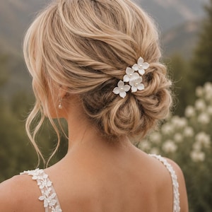 Può includere: Capelli biondi raccolti in uno chignon, adornati con tre forcine bianche a forma di fiore. Si vede la schiena della donna, con le spalle e il collo, e una spallina di pizzo del vestito.