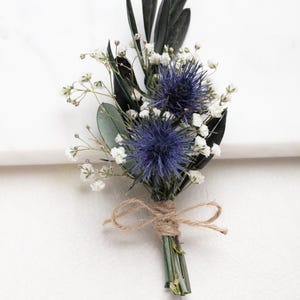 Könnte beinhalten: Handgefertigte Boutonniere mit leuchtend blauen Distelblüten, zartem weißen Schleierkraut und dunkelgrünen Olivenblättern. Die Stiele sind mit Bindfaden gebunden, was ein rustikales und elegantes Blumenarrangement ergibt.
