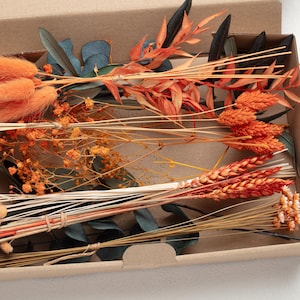 May include: A collection of dried flowers in a cardboard box. The arrangement includes orange bunny tails, wheat, and other dried stems in shades of orange, brown, and green. The box is open, revealing the contents.