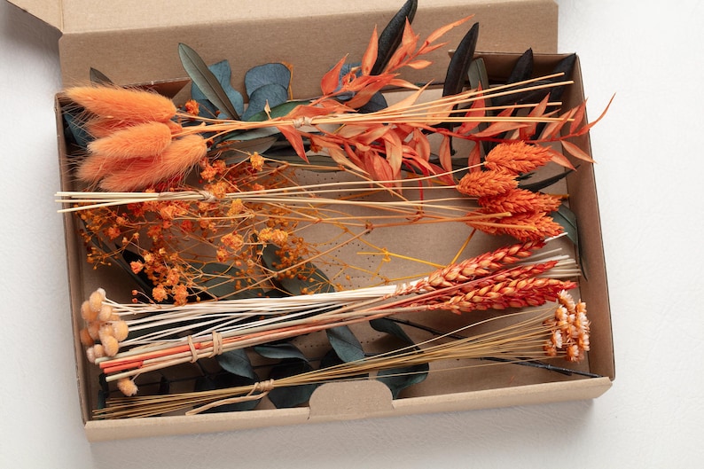 May include: A collection of dried flowers in a cardboard box. The arrangement includes orange bunny tails, wheat, and other dried stems in shades of orange, brown, and green. The box is open, revealing the contents.