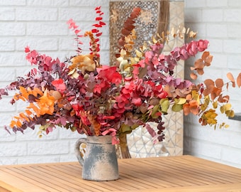 May include: A bouquet of dried eucalyptus branches in shades of red, orange, and yellow arranged in a grey ceramic pitcher on a wooden table.