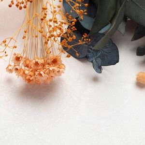 May include: Close-up of dried floral arrangement with orange, peach, and green elements. The composition includes cone-shaped orange flowers, delicate peach-colored blooms, and dark green eucalyptus leaves, all against a textured white background.