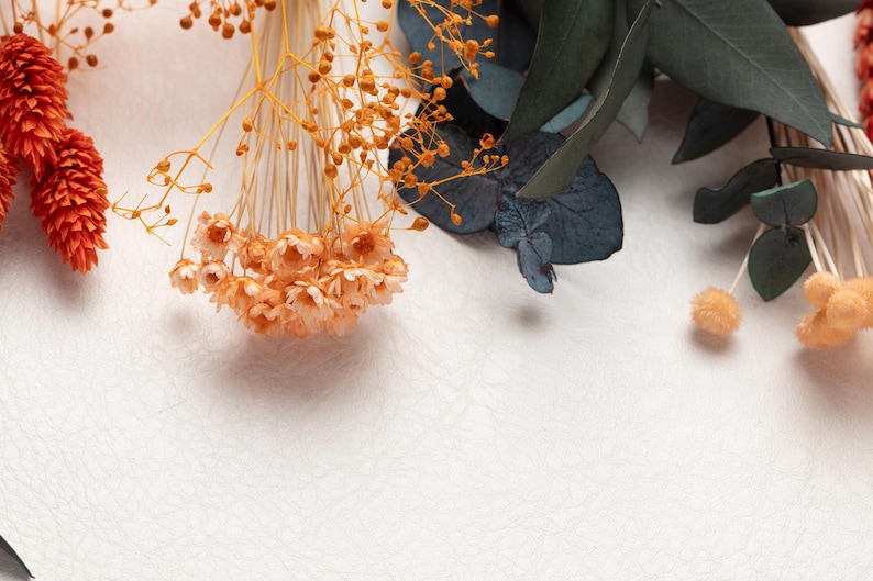 May include: Close-up of dried floral arrangement with orange, peach, and green elements. The composition includes cone-shaped orange flowers, delicate peach-colored blooms, and dark green eucalyptus leaves, all against a textured white background.