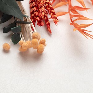 May include: An overhead shot of dried floral elements in shades of orange, red, and green against a white background. The arrangement includes fluffy orange pom-poms, spiky orange leaves, and dark green eucalyptus leaves.