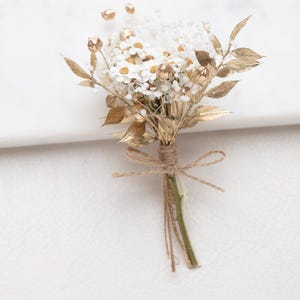 Könnte beinhalten: Ein kleiner Strauß weißer Blumen mit goldfarbenen Akzenten und Blättern, gebunden mit Bindfaden. Das Arrangement zeigt kleine weiße Blüten und goldfarbenes Laub, wodurch eine elegante und natürliche Ästhetik entsteht. Geeignet für Hochzeiten oder besondere Anlässe.