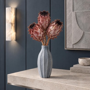 Pode incluir: Um vaso de cerâmica cinzenta com textura canelada vertical contém três flores de protea secas. As flores apresentam pétalas pontiagudas em camadas em tons de rosa suave, castanho e vermelho escuro, com centros escuros e felpudos.