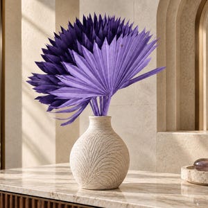 Könnte beinhalten: Eine runde, cremeweiße Keramikvase mit einer strukturierten, geschnitzten Blattverzierung auf der Oberfläche. In der Vase stecken mehrere leuchtend tiefviolette, getrocknete Palmblätter, die fächerförmig angeordnet sind und einen Kontrast bilden.